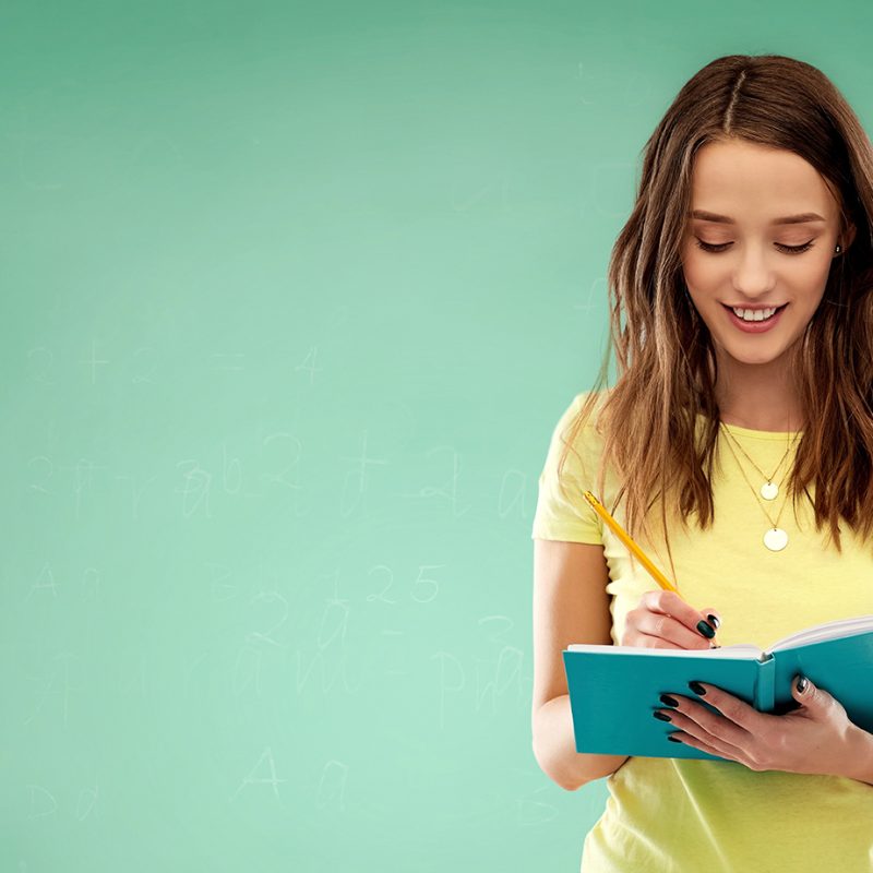 education, school, inspiration and people concept - smiling teenage student girl in yellow t-shirt writing to diary or notebook by pencil over green chalk board background