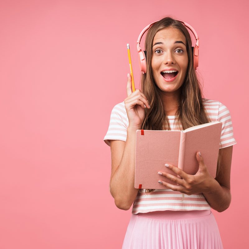 Image of excited beautiful girl writing down notes at diary book and listening to music with headphones isolated over pink background